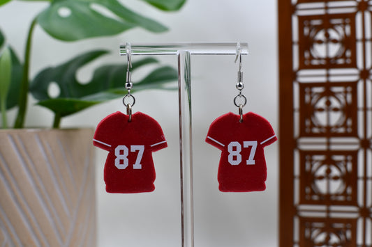 Red earrings shaped like miniature jerseys with the number 87 on a stand.